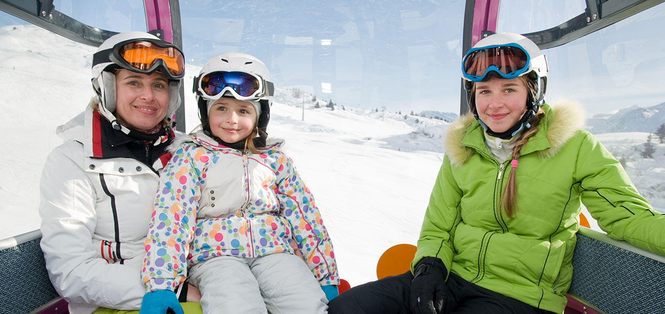 Mamma con due figlie in tuta da sci dentro funivia con paesaggio innevato sullo sfondo