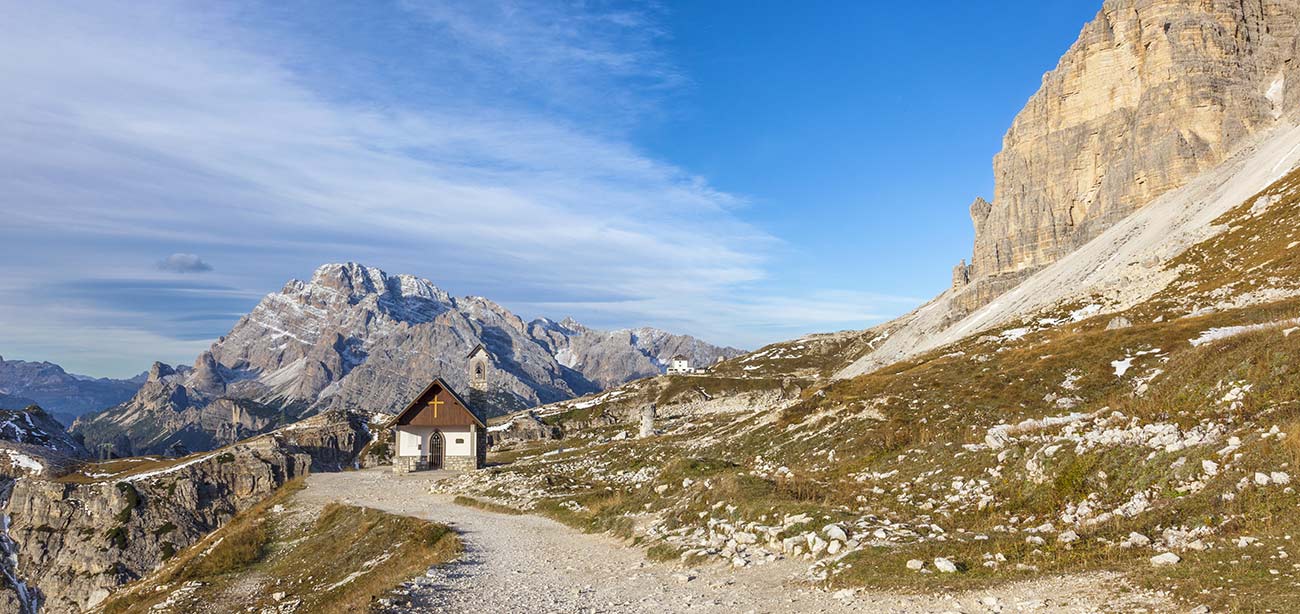 Sentiero sterrato porta a casetta alpina con Dolomiti e cielo azzurro sullo sfondo